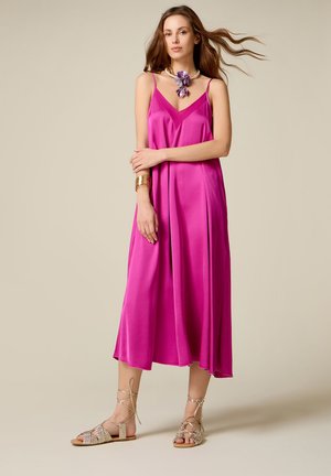 Frau in einem ärmellosen, knallpinkfarbenen Midikleid, die Schnürsandalen, eine Blumenkette und ein goldenes Armband vor einem beigen Hintergrund trägt.