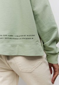 Sudadera verde claro de corte relajado, con un print de logo en el dobladillo. Se combina con pantalones texturizados de color crema.