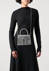 Sac à main en argent embossé crocodile avec une forme structurée, des poignées courtes et un détail de nœud décoratif, porté sur une robe noire.