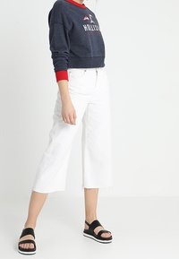 Námořnická zkrácená mikina s červenými detaily, bílé široké džíny s třásněmi na spodním lemu a černé sandály s bílou podešví.