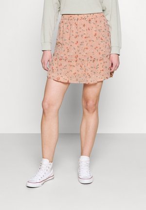 Personne portant une mini-jupe fleurie rose avec un ourlet à volants, un top vert clair à manches longues et des baskets montantes blanches, debout devant un fond blanc.