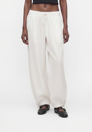 Femme portant un pantalon blanc ample avec cordon de serrage et poignets élastiques ainsi que des chaussures Mary Jane noires sur fond blanc uni.