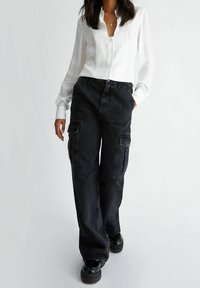 Pantaloni cargo neri con tasche laterali, abbinati a una camicia bianca con bottoni. Il tessuto appare texture, e le scarpe sono nere lucide.