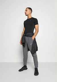 Schwarzes, eng anliegendes kurzärmeliges Hemd, graue Sporthose mit Seitentaschen und schwarze Turnschuhe. Eine leichte Jacke um die Taille gebunden.
