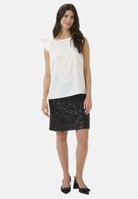 Femme portant un top sans manches blanc avec un nœud sur l'épaule, une jupe noire à sequins arrivant au genou et des escarpins noirs à talons ouverts, se tenant devant un fond blanc.