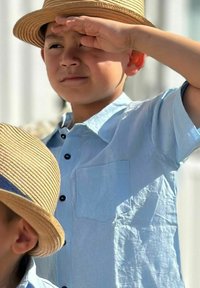 Vaaleansininen lyhytnhihainen paita mustilla napilla ja rintataskulla, käytetty vaaleanruskean olkihatun alla, sileä tekstuuri ja rento istuvuus.