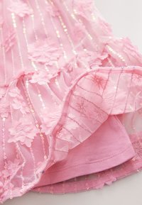 Rožnata prosojna tkanina z rožnatimi aplikacijami in navpičnimi bleščicami, slojevito prek trdne rožnate podlage, prikazuje podrobnosti šivov in teksture.