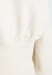 Gros plan sur un pull en tricot couleur crème mettant en valeur des détails de tissu froncé à la taille et sur une partie de la manche.