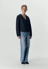 Mulher com cabelo encaracolado, vestindo uma jaqueta preta acolchoada, jeans azul claros e sapatos pretos, de pé contra um fundo claro e liso.