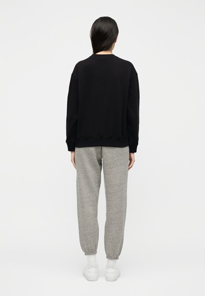 Άτομο με μακριά σκουρόχρωμα μαλλιά που φοράει μαύρο φούτερ, γκρι φόρμα, λευκές κάλτσες και λευκά αθλητικά, να στέκεται με την πλάτη προς τα εμπρός σε λευκό φόντο.