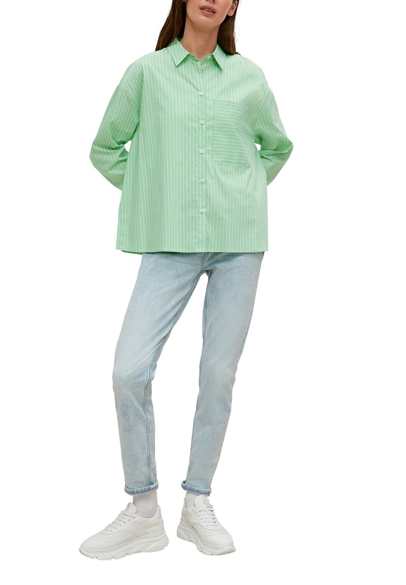 Lichtgroene overhem shirt met verticale strepen, enkele borstzak, losse pasvorm; gecombineerd met lichtblauwe spijkerbroek en witte sneakers.