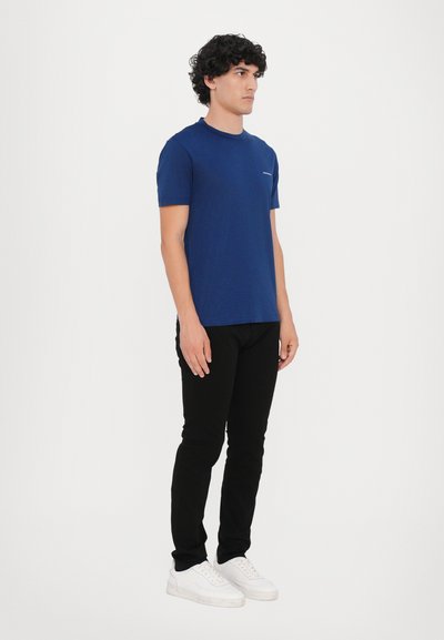 Tamsiai mėlyna trumpomis rankovėmis marškinėliai su apvaliu iškirpimu, dėvimi su juodomis siaurais kelnėmis ir baltomis sportinėmis bateliais, stovintys prieš paprastą baltą foną.