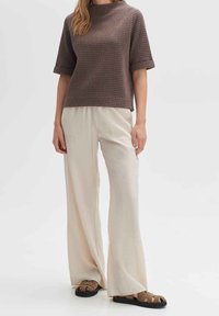 Top corto a maniche corte di colore marrone con texture abbinato a pantaloni a gamba larga beige chiaro. Modella indossa sandali alla schiava marroni e neri su uno sfondo neutro.