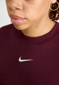 Burgundinis sportinis megztinis su gipiūro apykakle, su baltu Nike logotipu ant krūtinės. Lygus paviršius, artimas modelio veido kadras.