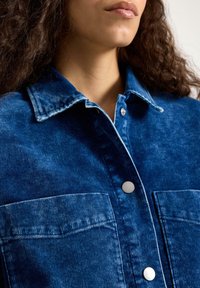 Džínová bunda tmavě modré barvy s dvěma předními kapsami, zapínáním na knoflíky a límečkem. Materiál má jemnou texturu a jemné známky opotřebení.