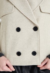 Vest dublu din lână amestecată, bej, cu nasturi negri și buzunare mari cu clapă. Textură netedă și design croit.
