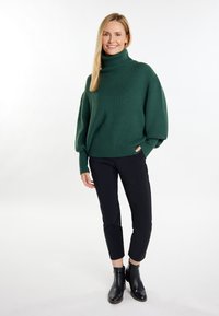 Pulover gros verde, tricotat, cu guler înalt, mâneci relaxate, asortat cu pantaloni negri evazați și cizme până la gleznă. Textură fină și model în relief.