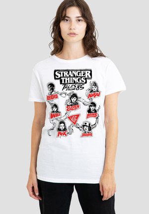 Young woman wearing white Stranger Things graphic tee with black-and-white character sketches and red names against a plain gray background.