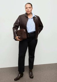 Brązowa skórzana kurtka, jasnoniebieska koszula z kołnierzykiem, czarne spodnie, brązowe buty, trzymający brązową torbę o fakturze, na jasnym tle.