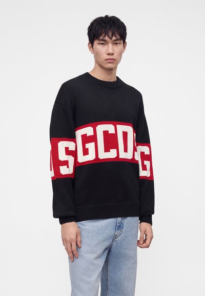 Jeune homme aux cheveux noirs portant un pull noir avec des lettres GCDS rouges et blanches sur la poitrine et un jean bleu clair.