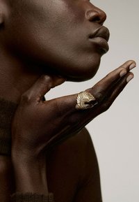 Anillo de oro con un diseño texturizado en forma de concha que se lleva en la mano. El tono de piel es oscuro, con la mano descansando suavemente cerca de la barbilla.