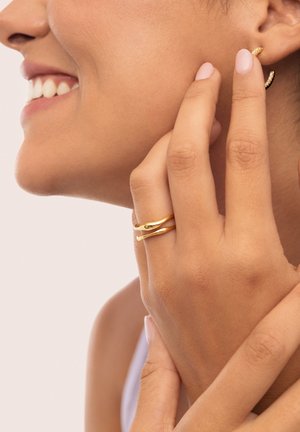 Anillos de metal dorado con un diseño retorcido, usados en los dedos. Pequeños pendientes de aro dorados con una superficie texturizada. Tonalidad de piel visible junto a la joyería.