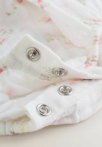 Belo blago z majhnimi roza in zelenimi cvetličnimi vzorci, opremljeno s srebrnimi pritisnimi gumbi in nabrani robovi za teksturiran zaključek.