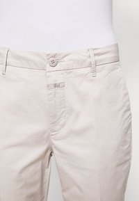 Pantalons beige clair avec une fermeture à bouton, une poche avant unique et une petite étiquette de logo. Le matériau semble lisse avec un léger brillant.