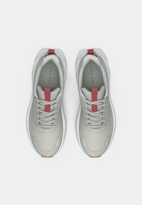 Gaiši pelēki sporta apavi ar teksturētu augšdaļu, sarkaniem akcentiem un baltu zoli. Iezīmes ietver šņorītes un uz zolītes iespiestu logotipu.