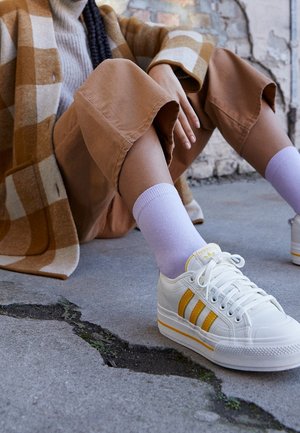 Białe sneakersy z żółtymi paskami, z teksturowaną gumową podeszwą, noszone z jasnofioletowymi skarpetkami, w połączeniu z brązową kwadratową marynarką i beżowymi szortami.