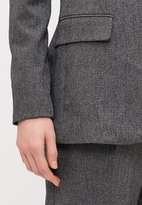Primo piano di un blazer grigio su misura, con tessuto texturizzato e con leggere macchie, colletto a scollo a V e una tasca sul petto.