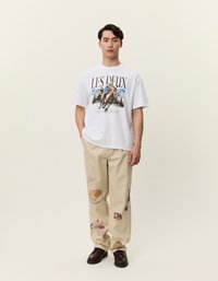 Biała bawełniana koszulka z nadrukiem górskiego konia, w połączeniu z beżowymi spodniami z ilustracjami związanymi z jedzeniem oraz brązowymi butami. Proste tło.