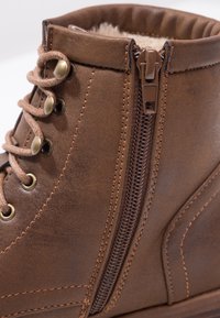 Brown leather boot featuring a textured finish, rounded toe, decorative stitching, brass eyelets, laces, and a side zipper for easy access.