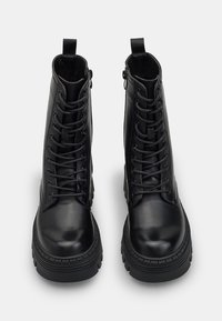 Botines de cuero negro con una textura suave, con cordones en la parte delantera y cremalleras laterales, que cuentan con una suela de goma gruesa y un tacón reforzado.