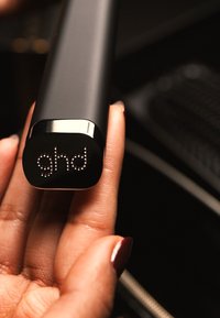 Prostownica do włosów w czarnym kolorze o gładkim matowym wykończeniu. Zawiera metaliczne akcenty i logo LED. Trzymana w ręce dla pokazania skali.