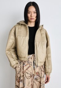 Beige quilted puffer jacket with a hood, featuring textured panels and a front zipper. Paired with a patterned beige skirt.