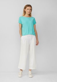 Vrouw staand tegen een grijze achtergrond, gekleed in een turkoois T-shirt met korte mouwen, witte wijde broek en witte sneakers.