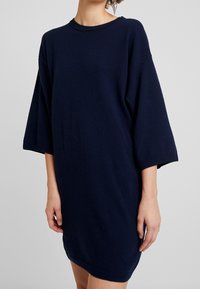 Granatowa, oversize'owa sukienka-sweterek z krótkimi rękawami, prążkowanym dekoltem i dołem. Wykonana z miękkiego materiału dzianinowego o luźnym kroju.