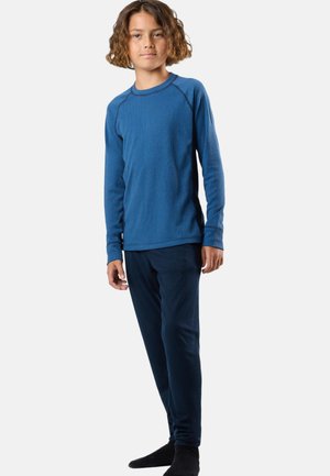 Child with curly hair wearing a blue long-sleeve shirt and dark blue pants, standing barefoot against a plain white background.