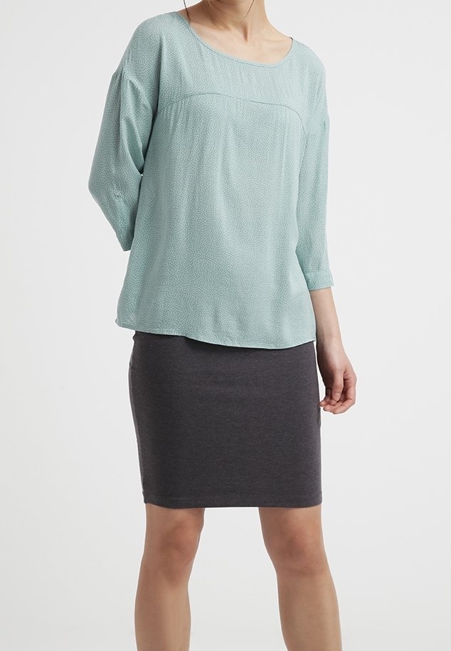 Femme portant une blouse ample bleu clair à manches longues et une jupe crayon gris foncé longueur genou sur un fond uni.