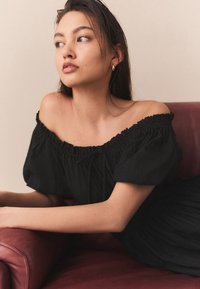 Schwarzes schulterfreies Oberteil mit Falten-Details, kurzen Puffärmeln und weicher Textur, kombiniert mit einem tiefroten Ledersessel.