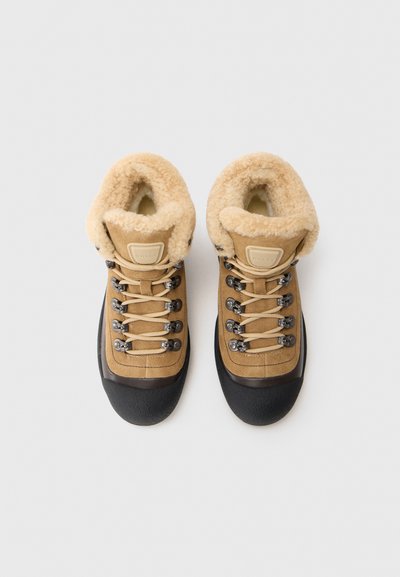 Botas de senderismo marrones con un suave cuello de felpa beige, ojales de metal y una puntera de goma negra. Diseño de ante texturizado y sistema de cordones en toda la bota.