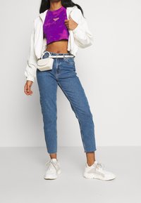 Sudadera blanca con cremallera, top corto morado, jeans de tiro alto azules y zapatillas blancas. Un pequeño bolso cruzado blanco acentúa el conjunto.