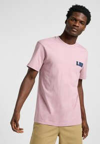 Camiseta de algodón de manga corta en rosa claro, con un logo de "LEE" en color azul marino en el lado izquierdo del pecho. Diseño simple, textura sin adornos.