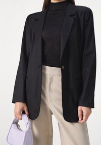Schwarzer Blazer mit langen Ärmeln, mit Reverskragen, einreihiger goldener Knopfverschluss, getragen über einem transparenten schwarzen Oberteil und beigen Hosen. Helles lila Handtasche.