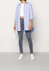 Camisa de rayas azul y blanca de gran tamaño, top corto blanco, jeans ajustados grises, zapatillas blancas, sosteniendo un pequeño bolso blanco.