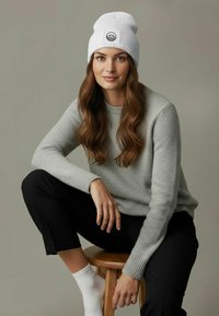 Gray knit sweater, white beanie with a mountain logo, black pants, and white socks, seated on a wooden stool with a neutral background.