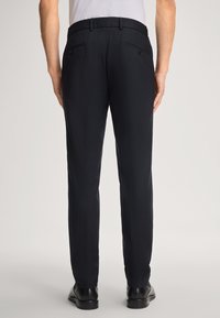 Achteraanzicht van een persoon in zwarte formele broek, zwarte nette schoenen en een wit ingestopt shirt tegen een effen achtergrond.