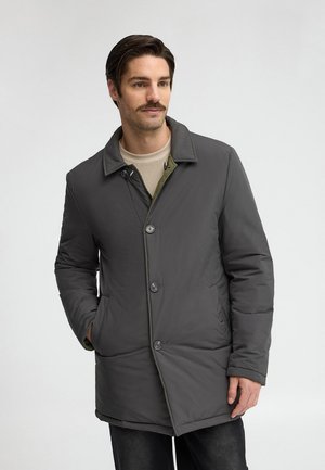 Hombre con bigote que lleva un abrigo acolchado gris oscuro sobre un suéter beige, de pie con una mano en el bolsillo contra un fondo liso.