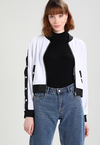 Kapat bomberjacka i vitt med svarta accenter, ribbade ärmslut och dragkedja. Bärs över en svart ribbad polotröja med högmidjade jeans.